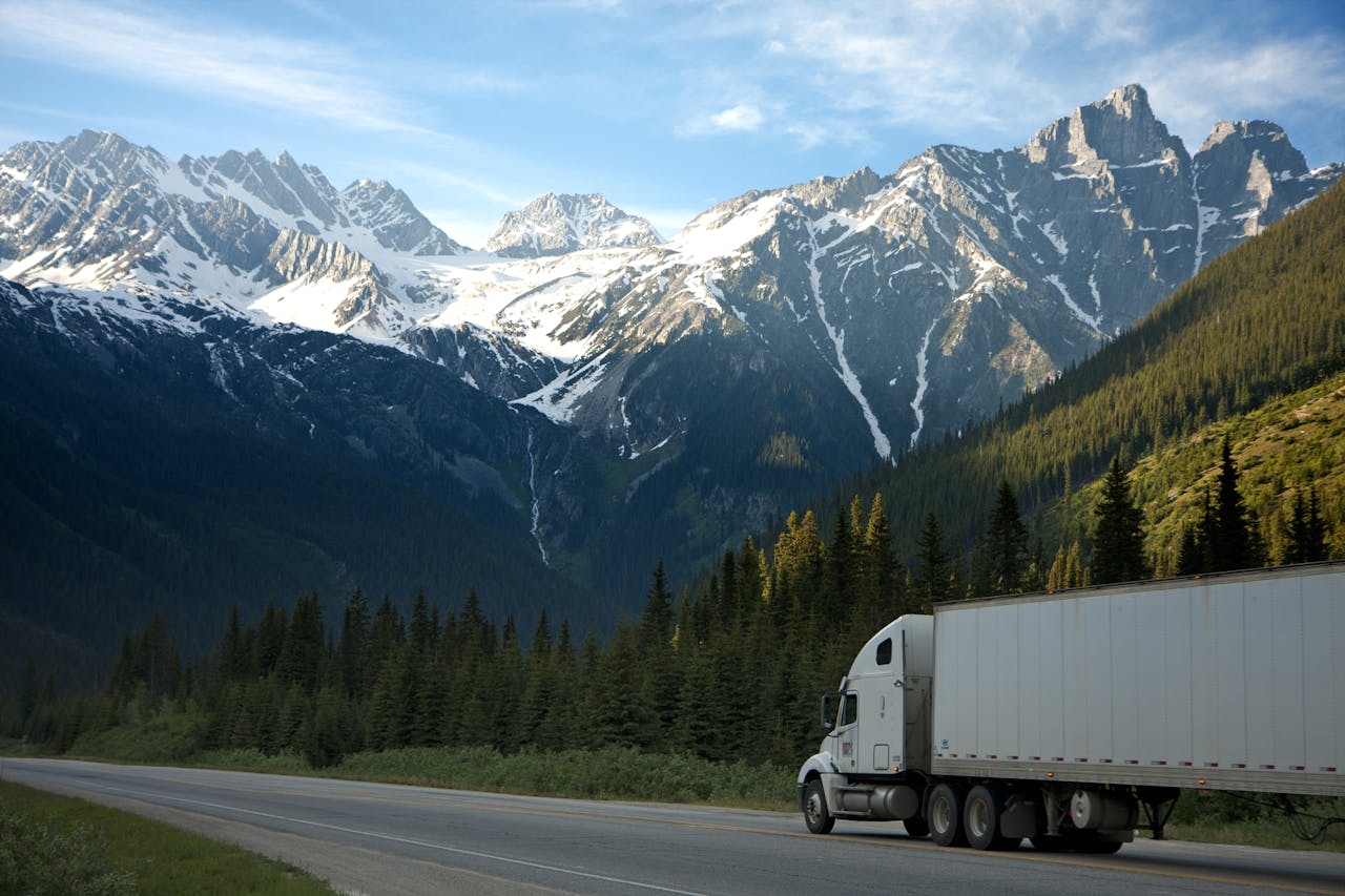 our-services-1 A semi-truck travels along a highway with snow-capped mountains in the background.